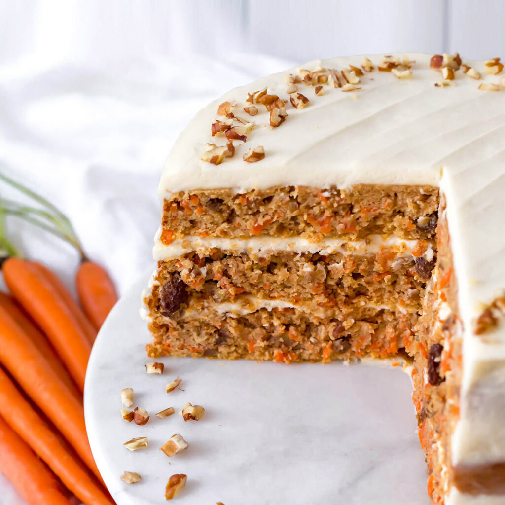 a frosted 3-layer carrot cake with a quarter of the cake cut out, on a marble cake stand with fresh carrots in the background