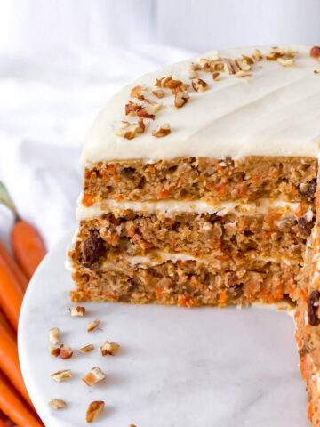 a frosted 3-layer carrot cake with a quarter of the cake cut out, on a marble cake stand with fresh carrots in the background