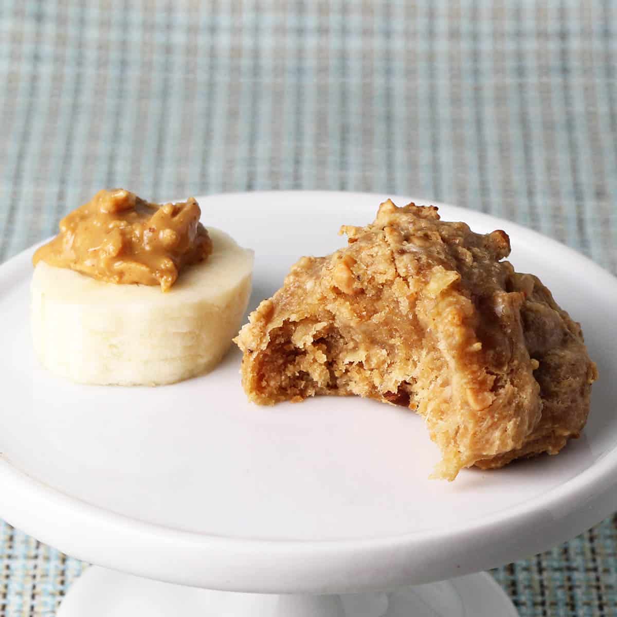 bitten chewy banana peanut butter protein cookie next to a slice of banana with peanut butter on top, on a white plate with a light blue textured background