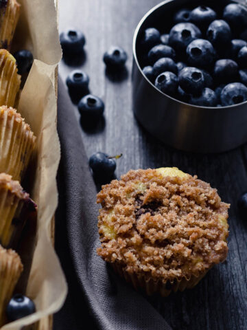 on the left, 5 small blueberry muffins in a small wooden box, and on the right, a muffin with textured crumble topping and a metal measuring cup filled with fresh blueberries, and a few blueberries out of the cup on a dark grey wooden surface