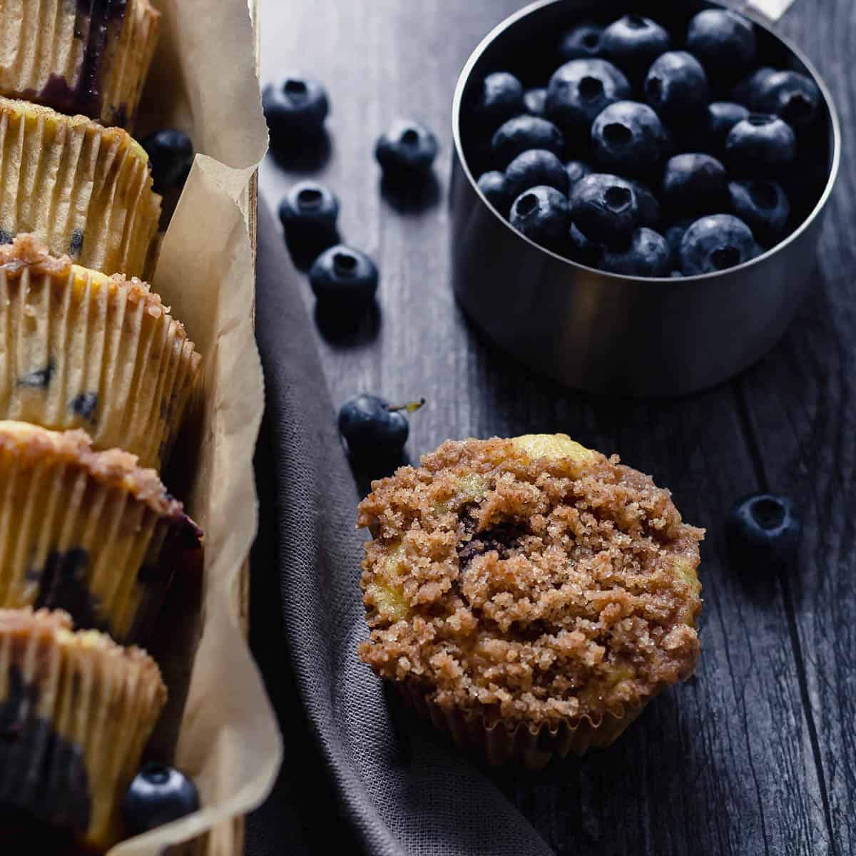 on the left, 5 small blueberry muffins in a small wooden box, and on the right, a muffin with textured crumble topping and a metal measuring cup filled with fresh blueberries, and a few blueberries out of the cup on a dark grey wooden surface