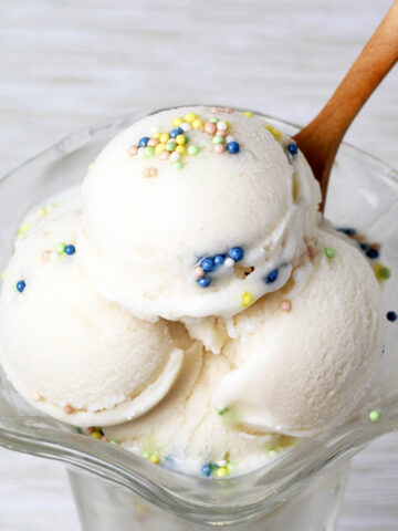 ice cream dish with vanilla looking (cake batter flavored) protein ice cream in rounded scoops, topped with nonpareil sprinkles and with a wooden dessert spoon