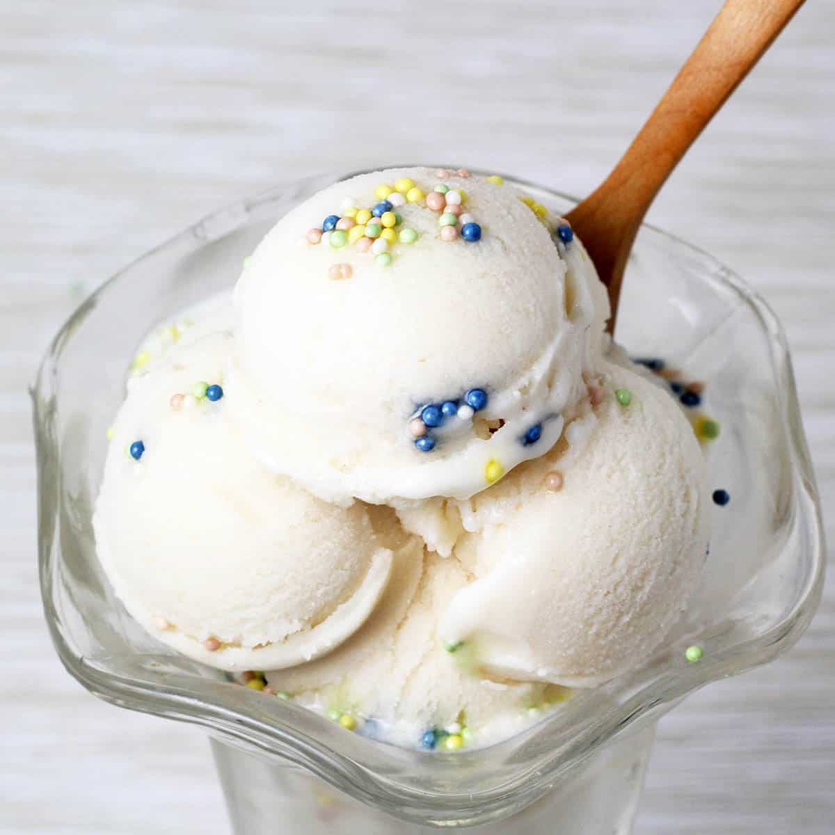 ice cream dish with vanilla looking (cake batter flavored) protein ice cream in rounded scoops, topped with nonpareil sprinkles and with a wooden dessert spoon