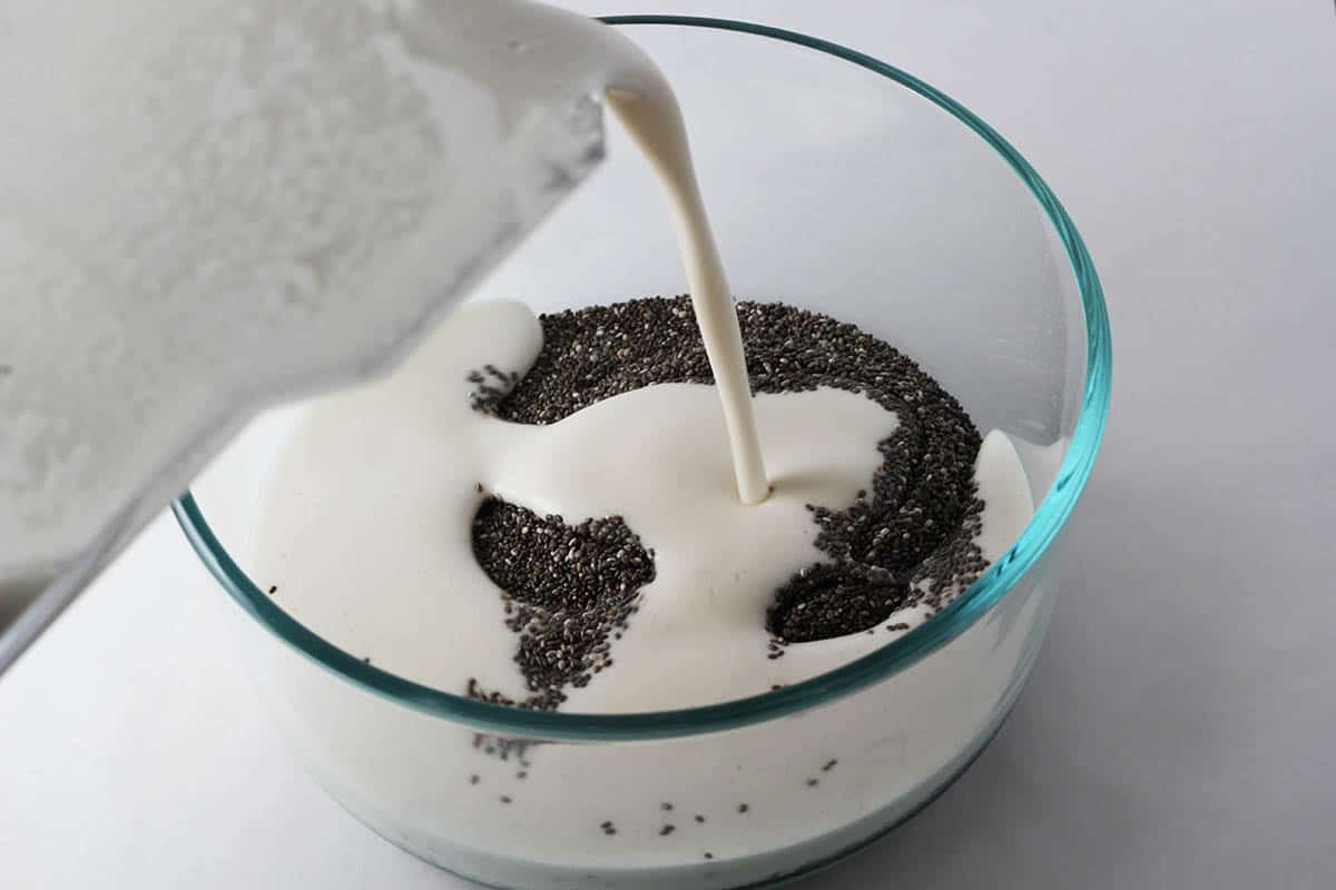 glass bowl with chia seeds and almond milk being poured in, on a white background