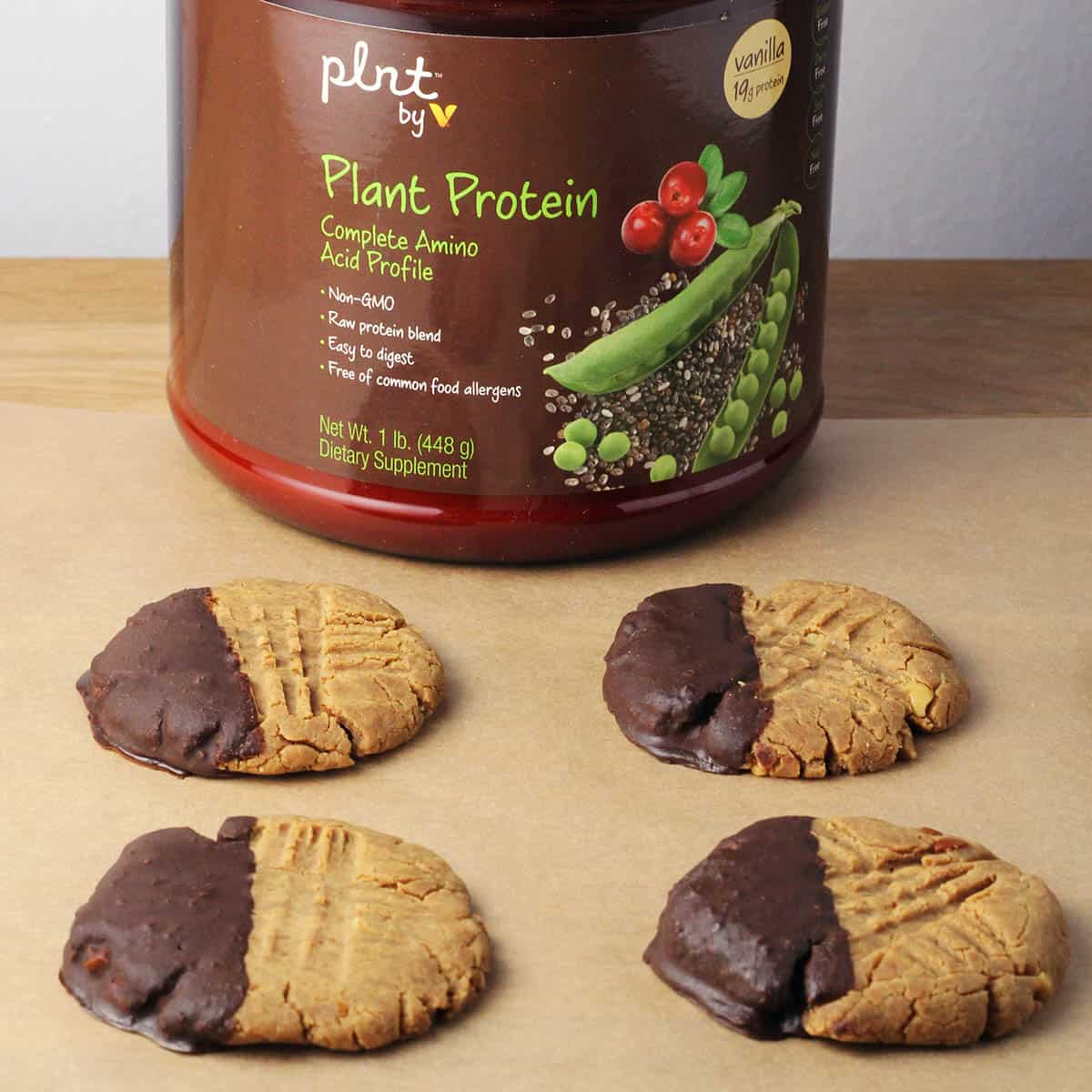four peanut butter protein cookies half dipped in chocolate in front of a canister of plnt brand protein powder