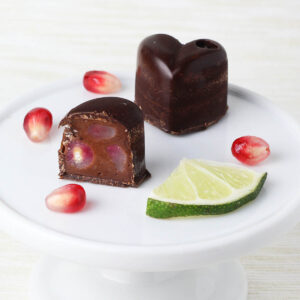 one and a half heart shaped filled chocolates with pomegranate seeds and a lime slice on a small white plate