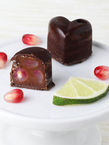 one and a half heart shaped filled chocolates with pomegranate seeds and a lime slice on a small white plate