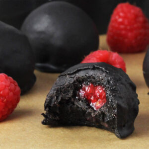 close up of a bitten very dark chocolate covered protein ball with raspberry center that looks heart shaped, with bright fresh raspberries and more protein balls behind it on a sheet of unbleached parchment paper