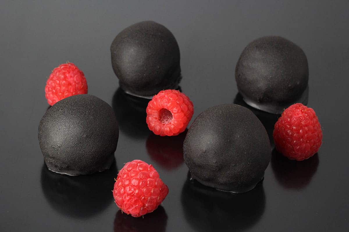 very dark chocolate covered protein balls with bright fresh raspberries on a black background