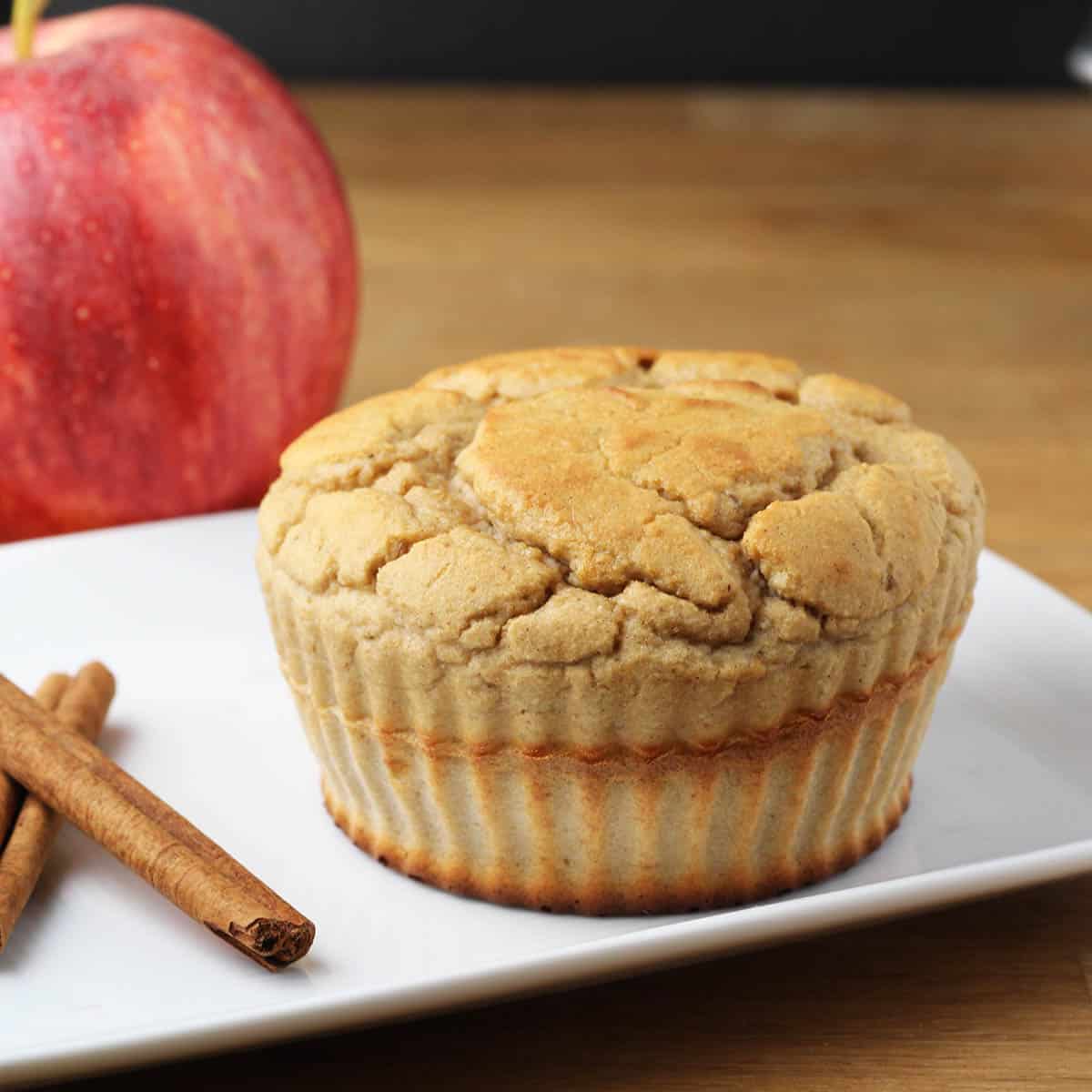 a cinnamon apple protein muffin on a white plate with two cinnamon sticks and an apple in the background on a wooden table