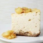 a straight on view of a slice of cheesecake that has cinnamon apples in a layer between the crust and cheesecake, on top of the slice, and in front of the slice on a white plate