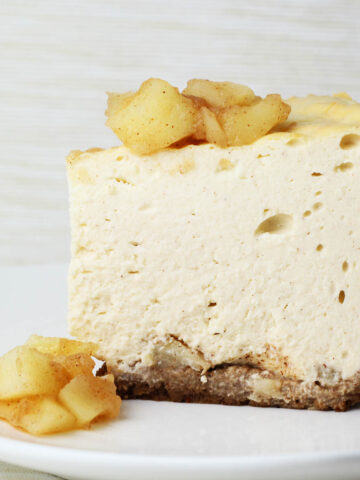 a straight on view of a slice of cheesecake that has cinnamon apples in a layer between the crust and cheesecake, on top of the slice, and in front of the slice on a white plate