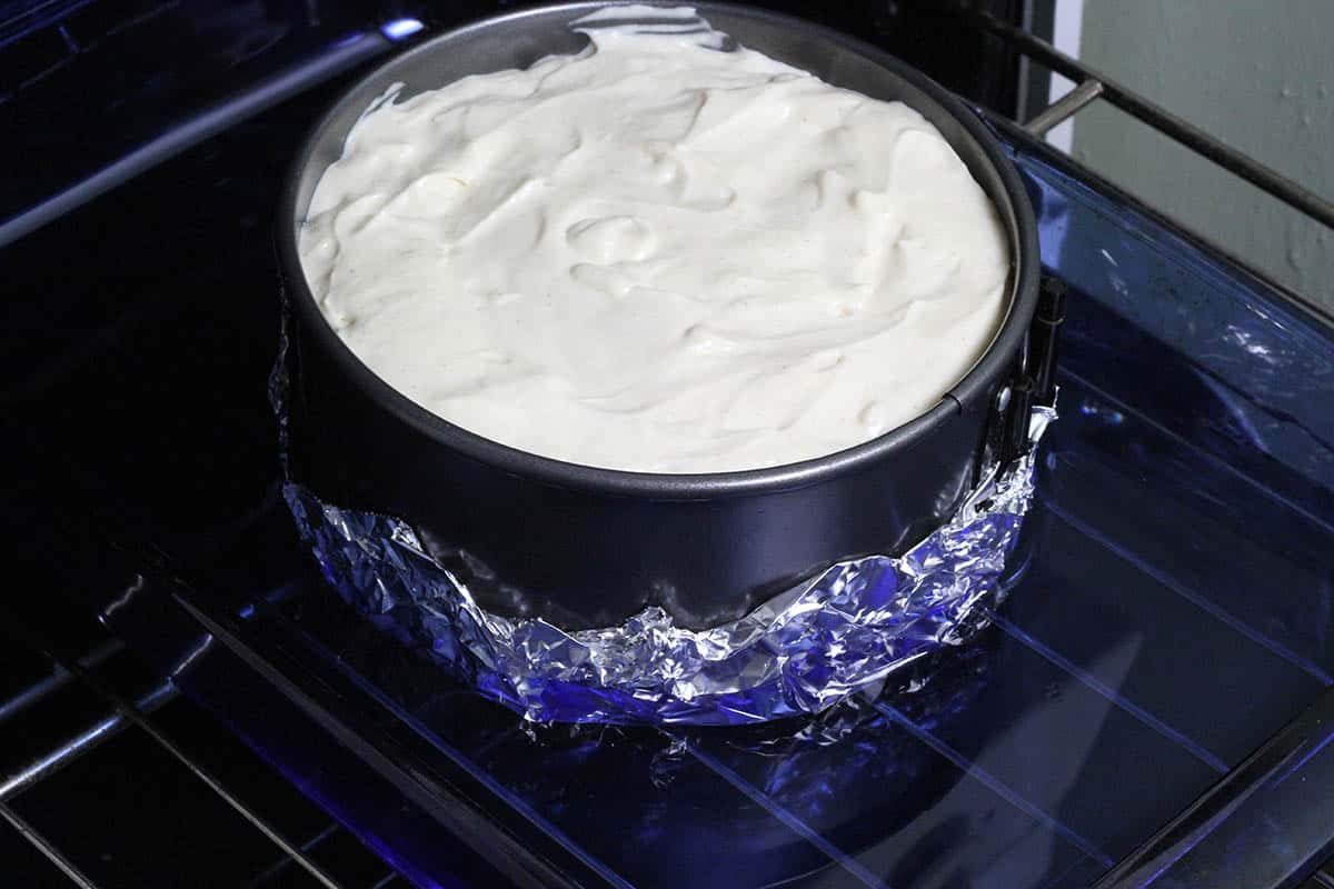 unbaked cheesecake in a springform pan that has foil around the bottom and in a pan of water (a water bath) on an oven rack