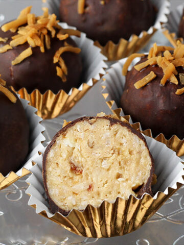 several full and one cut in half coconut caramel protein balls that are covered in chocolate and sprinkled with toasted coconut, each in a gold paper cup