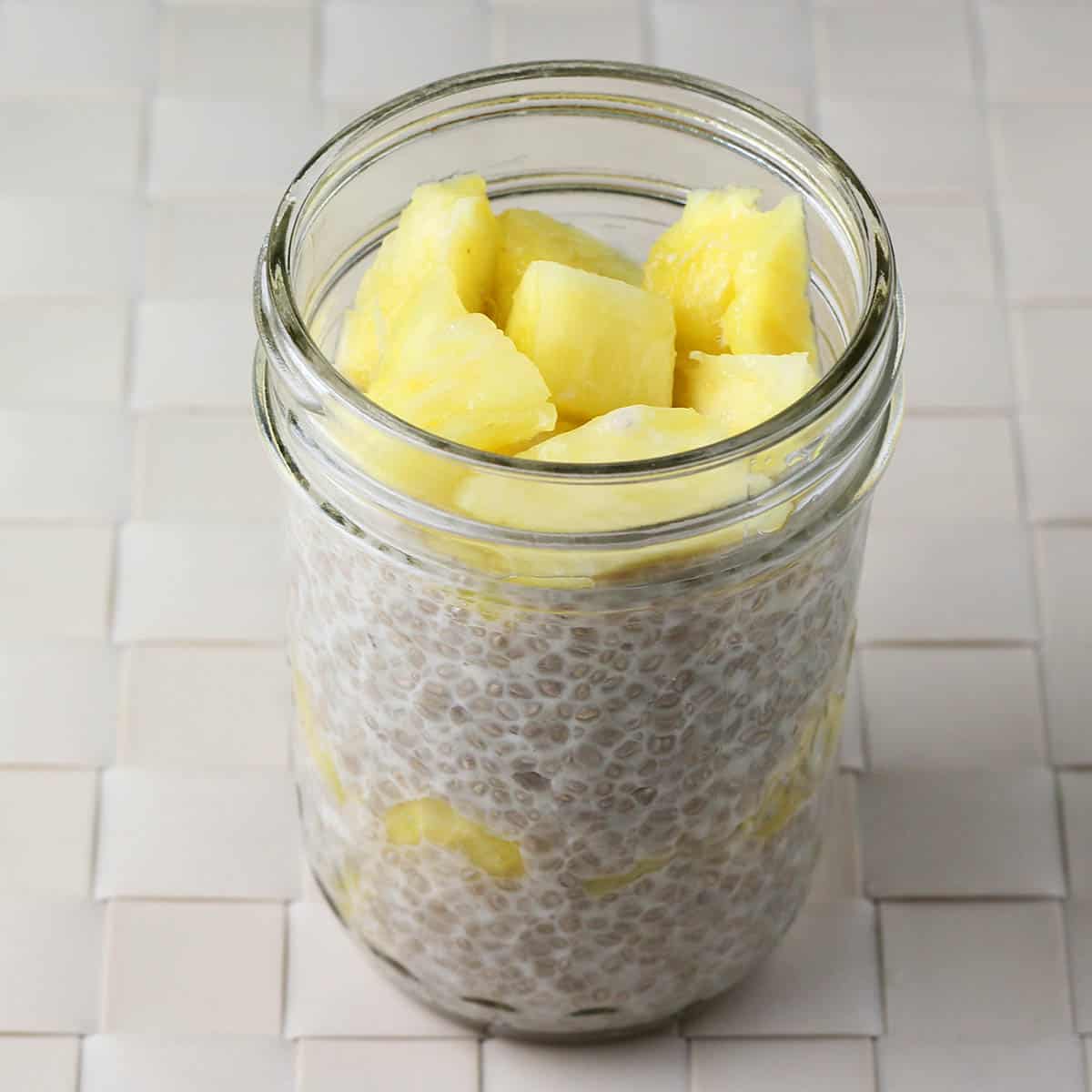 small round jar of coconut chia pudding layered with fresh pineapple on a woven beige placemat