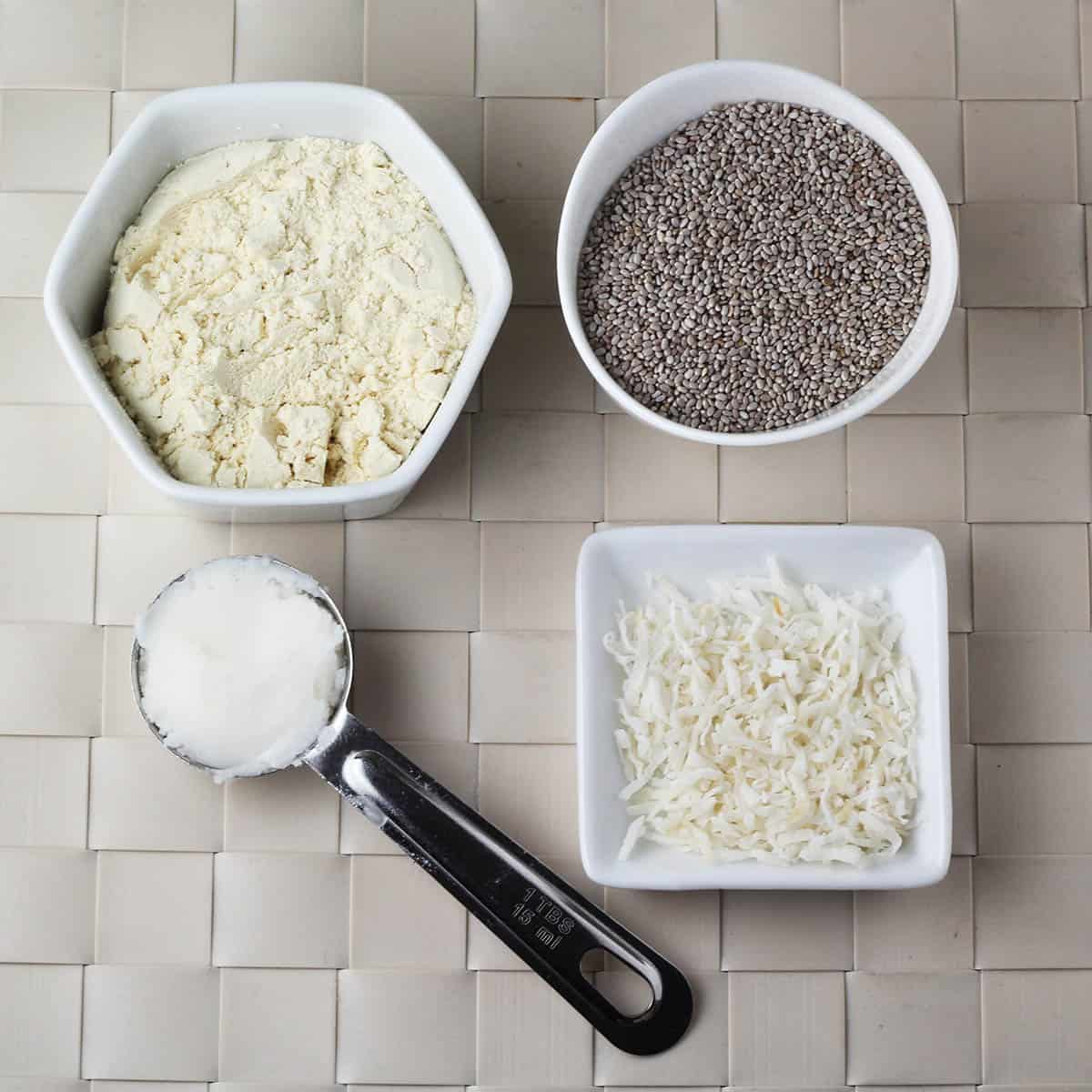 ingredients for coconut chia pudding: a tablespoon of coconut butter, a small dish of protein powder, a small dish of shredded coconut, and a small dish of chia seeds on a woven beige placemat