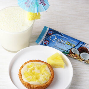 one coconut pineapple tartlet and a piece of fresh pineapple on a white plate with a quest bar and a coconut drink on a beige background