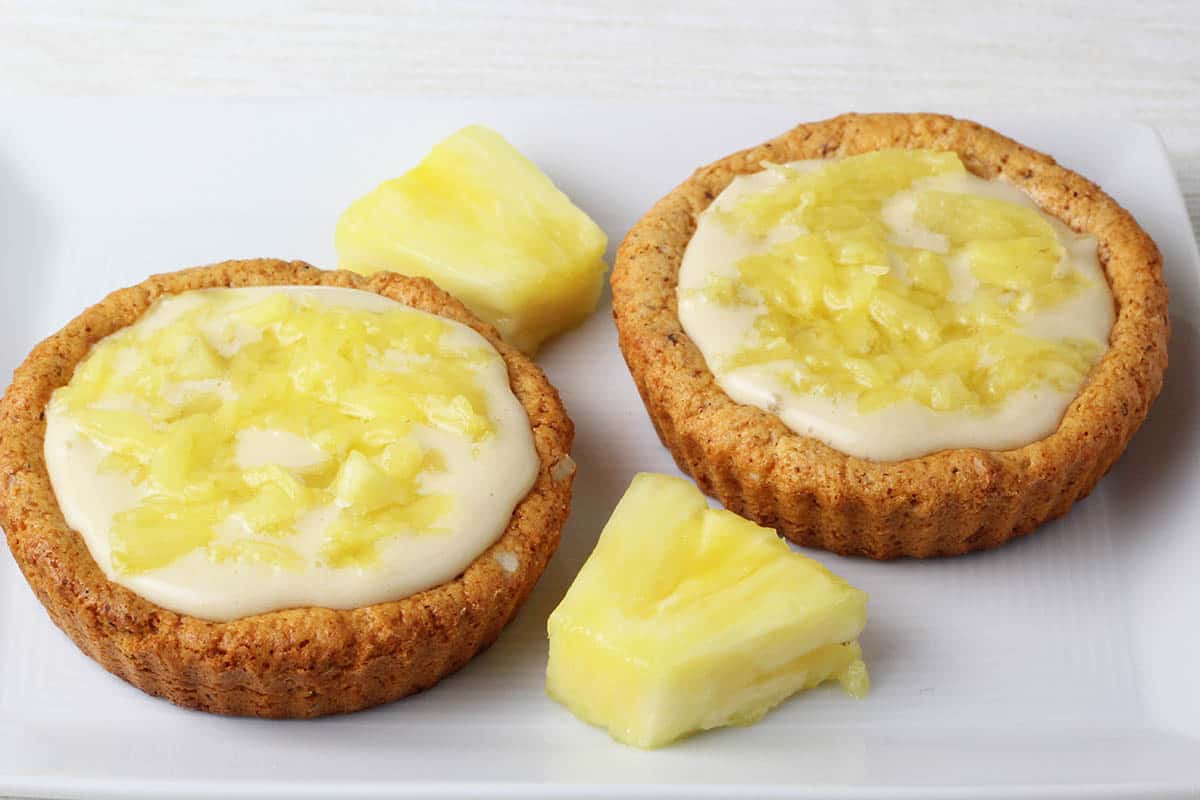 two mini tarts filled with vanilla whey cream filling and fresh pineapple, with two pieces of pineapple on a white plate