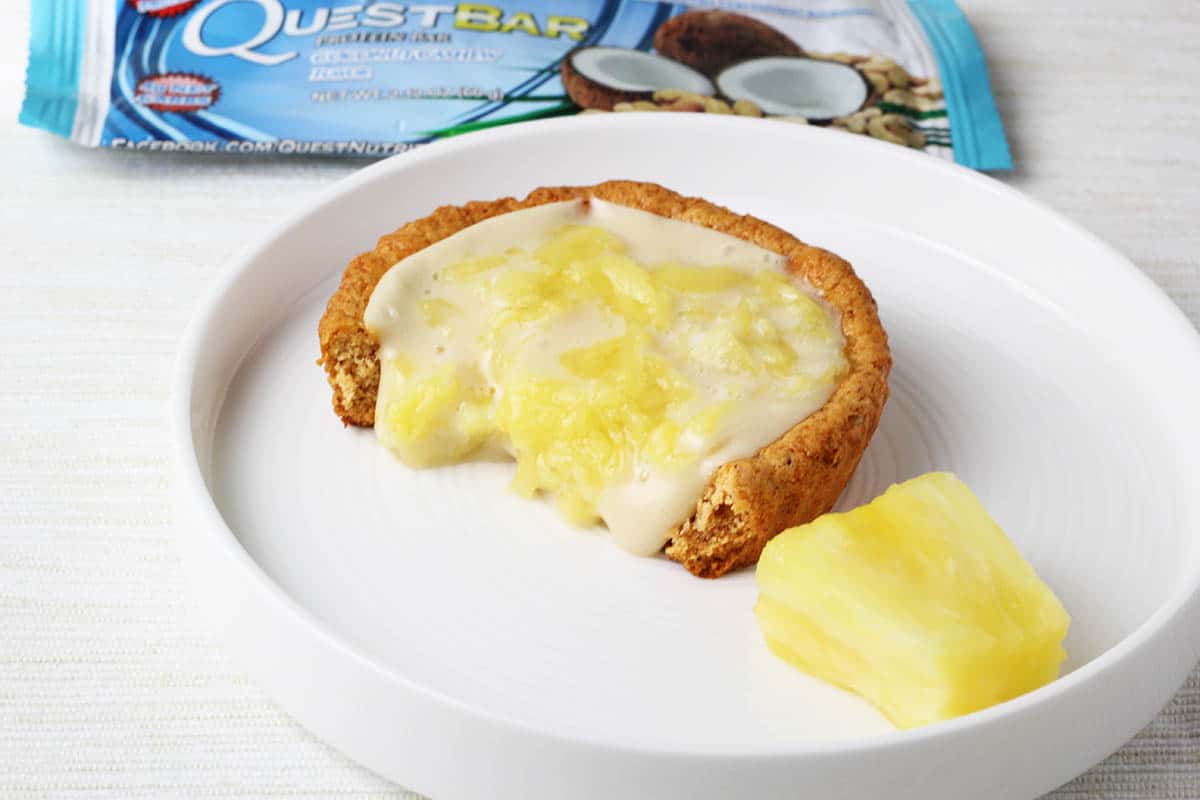 a bitten coconut pineapple tartlet on a white plate with a quest bar wrapper in the background