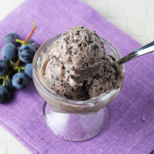 small glass dessert dish with a few scoops of grape gelato on a purple linen and with a small bunch of purple grapes