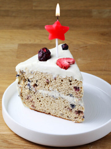 slice of confetti protein layer cake topped with freeze dried fruit with a lit red star candle on a white plate on a wooden table
