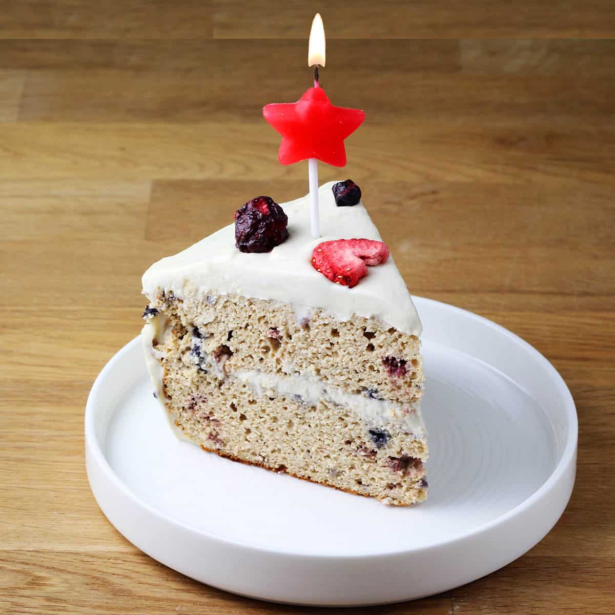 slice of confetti protein layer cake topped with freeze dried fruit with a lit red star candle on a white plate on a wooden table