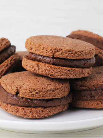 six chocolate sandwich protein cookies filled with thick chocolate protein frosting on a white plate with a light background