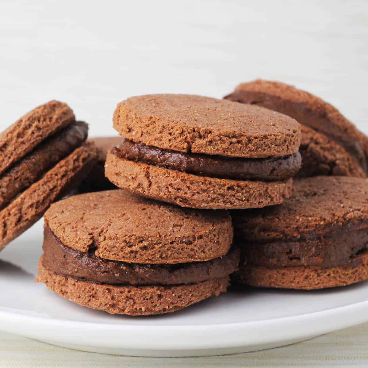 six chocolate sandwich protein cookies filled with thick chocolate protein frosting on a white plate with a light background