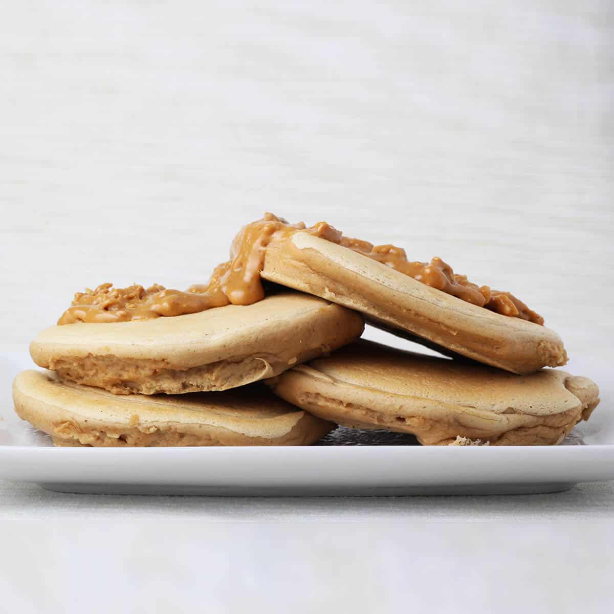 four fluffy peanut butter protein pancakes layered on a white plate and topped with crunchy peanut butter with a light backgorund