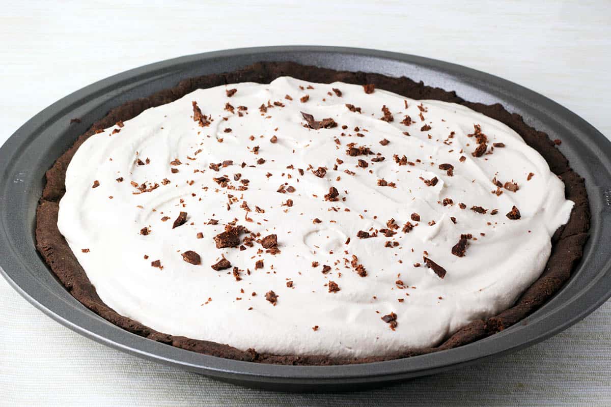 dark chocolate pie crust with filling and banana cashew whipped cream topping and chocolate shavings in a metal pie pan on a light background