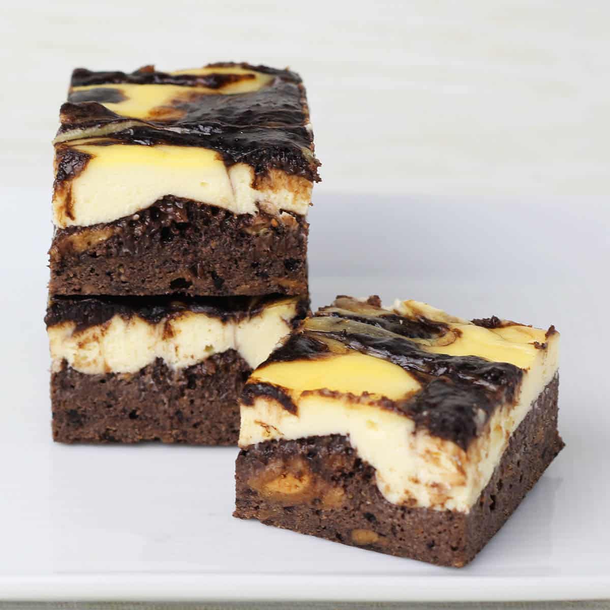 three pieces of cheesecake brownie (cheesecake layer on top of a brownie layer), two pieces are stacked on the left, and one is angled in front all on a white plate