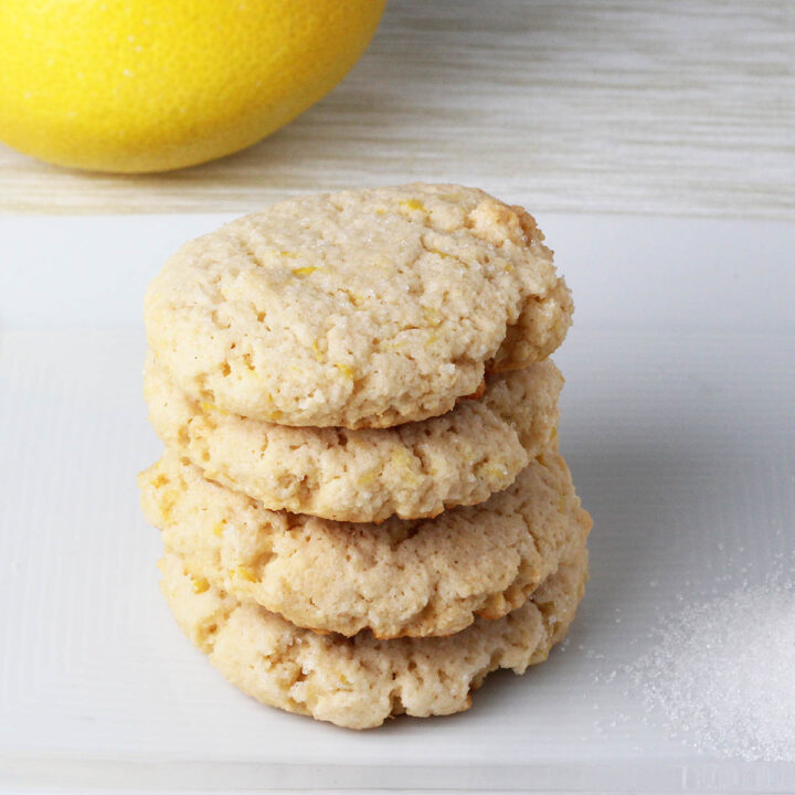 stack of four healthy lemon cookies on a white plate with granulated erythritol next to them and lemon in the background
