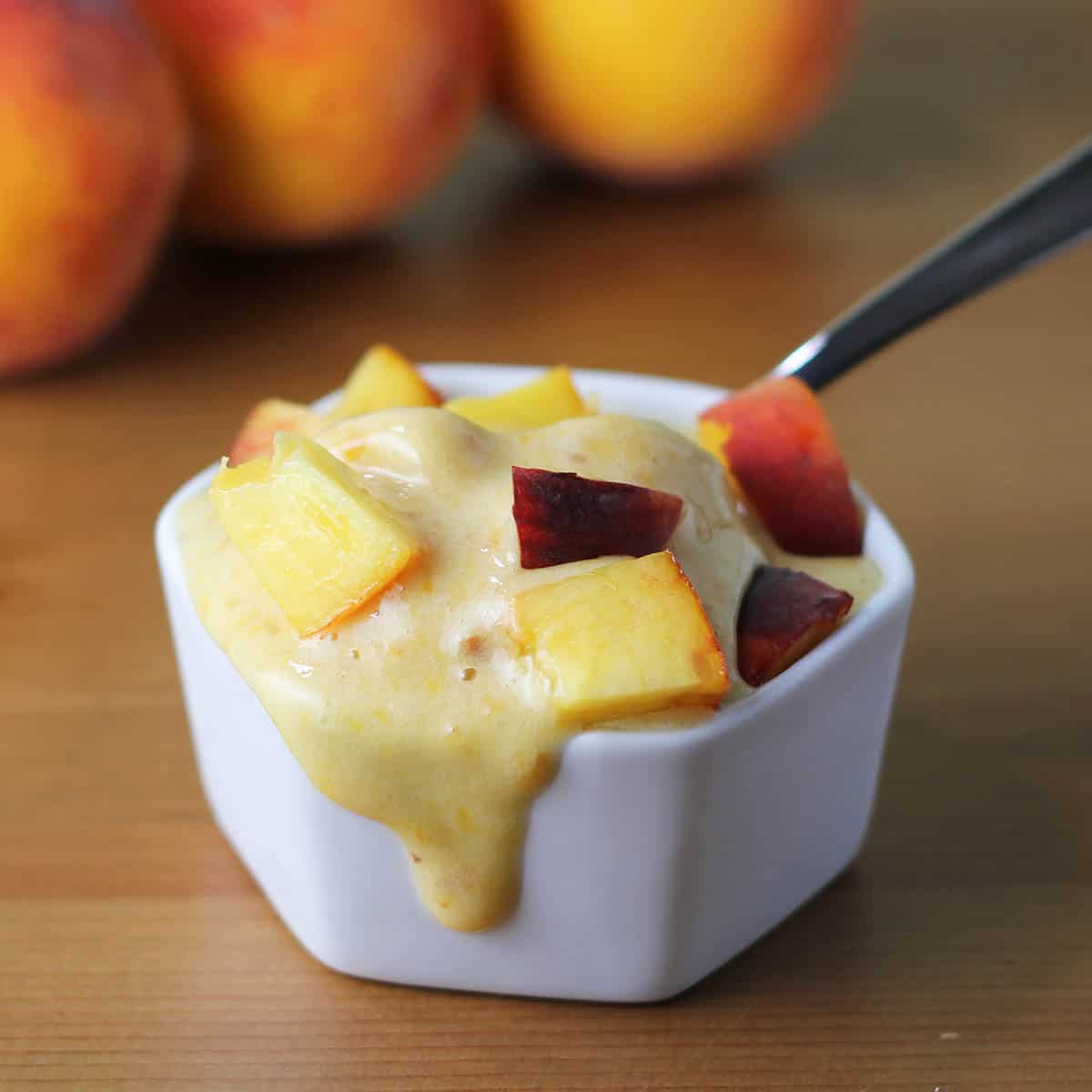 small white dish of melting peach ice cream with fresh cut peaches on top, a spoon in the dish, and three peaches in the background on a brown wooden table