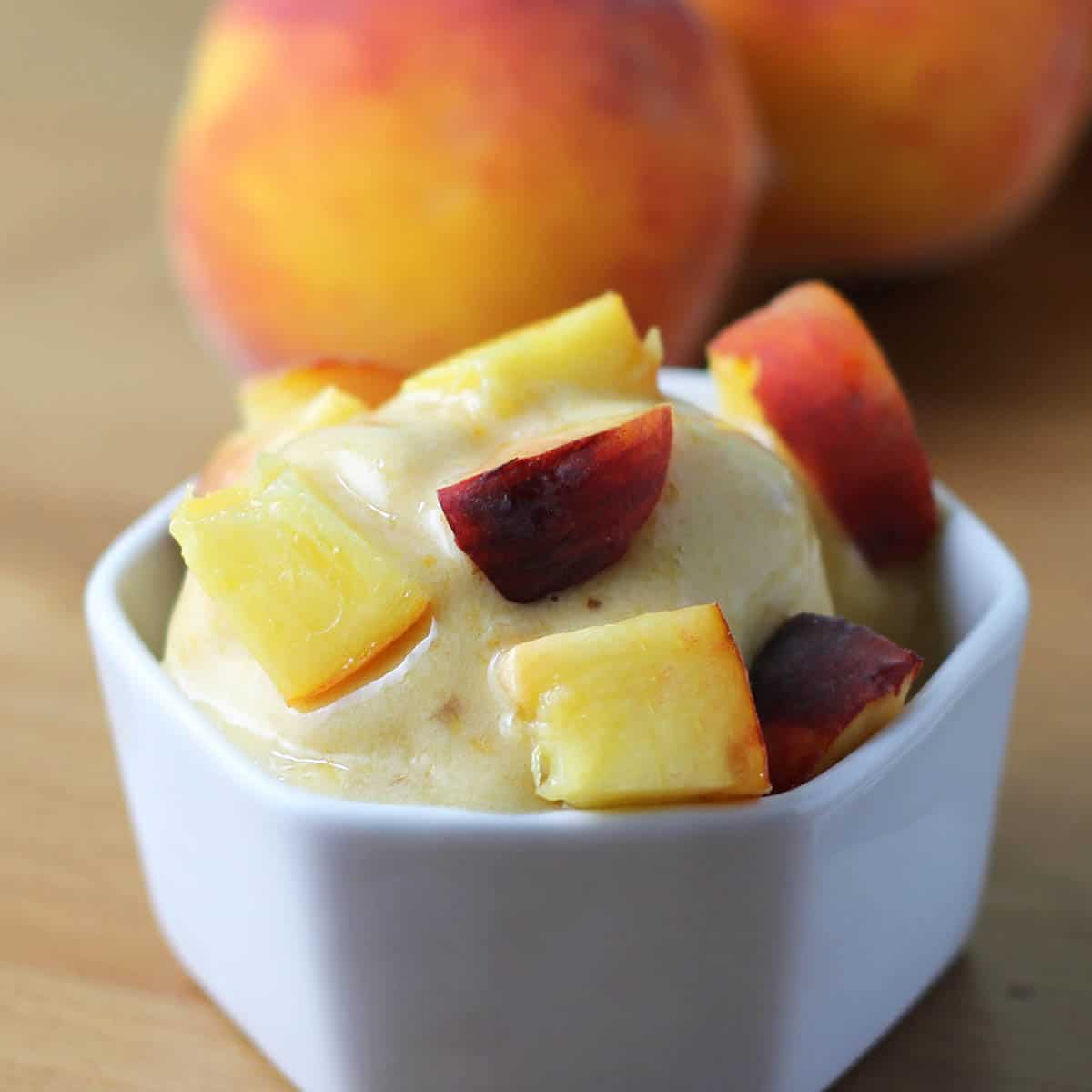 small white dish of peach ice cream with fresh cut peaches on top and two peaches in the background on a brown wooden table