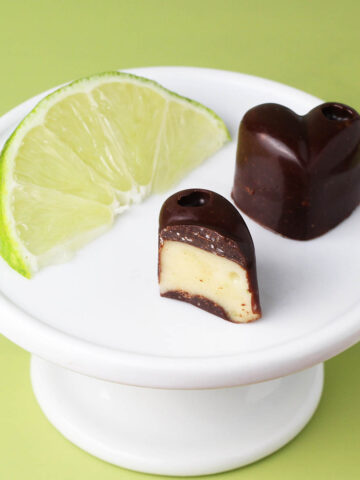 one full and one half piece of key lime filled chocolate hearts next to a slice of lime on a white plate on a lime green background