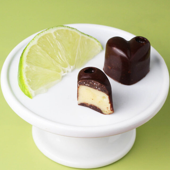 one full and one half piece of key lime filled chocolate hearts next to a slice of lime on a white plate on a lime green background