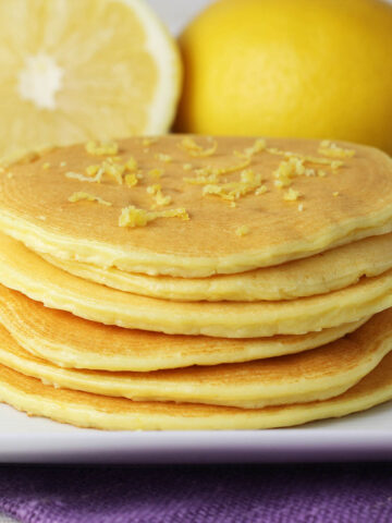 stack of 6 thin lemon ricotta pancakes on a white plate with one and a half lemons in the background and a purple linen napkin underneath