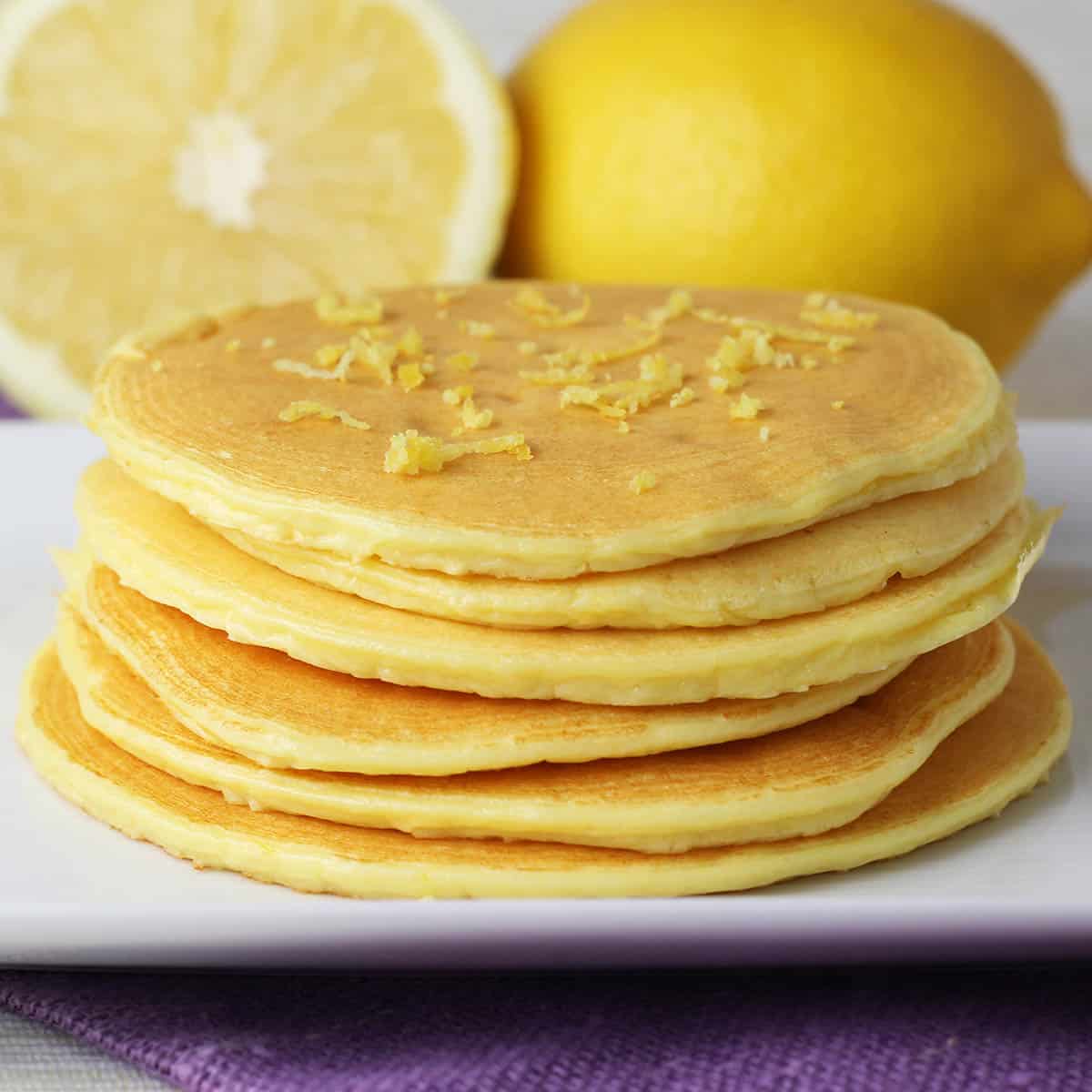 stack of 6 thin lemon ricotta pancakes on a white plate with one and a half lemons in the background and a purple linen napkin underneath