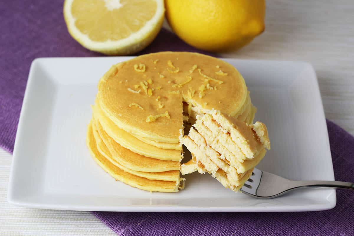stack of 6 thin lemon ricotta pancakes with a forkful being pulled out, on a white plate with one and a half lemons in the background and a purple linen napkin underneath