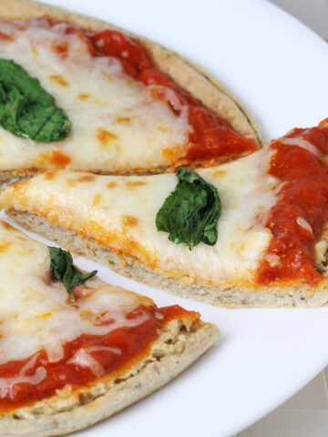 a slice of low carb pizza being pulled out of the whole pie, with basil on top, on a white plate