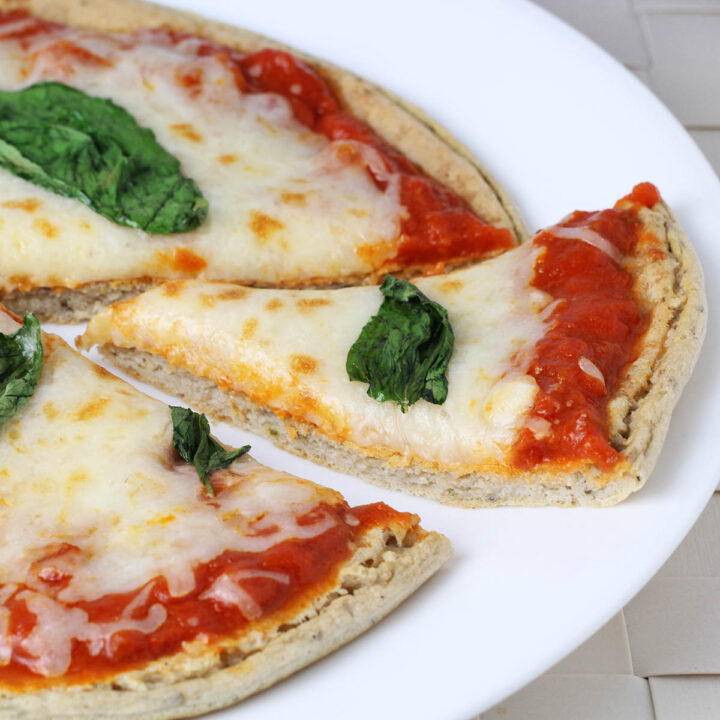 a slice of low carb pizza being pulled out of the whole pie, with basil on top, on a white plate