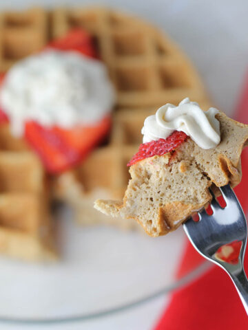 a bite of low carb protein waffle with strawberry and whipped cream on a fork in the front, and in the background is the rest of the waffle on a glass plate with a red linen napkin