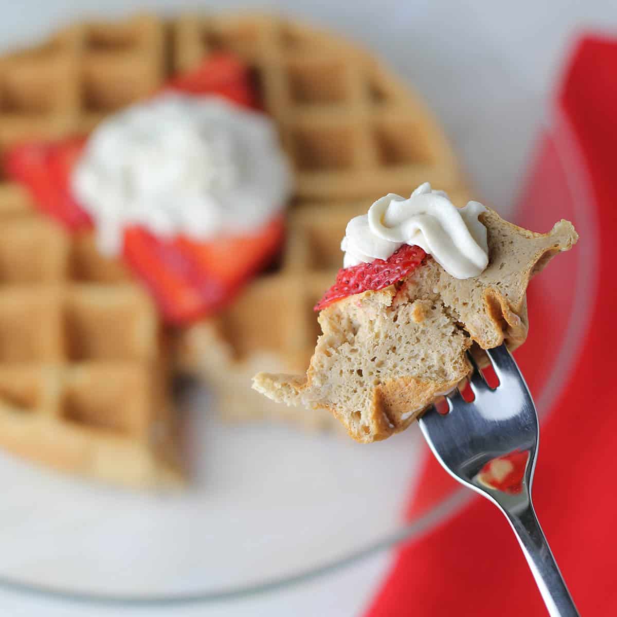 a bite of low carb protein waffle with strawberry and whipped cream on a fork in the front, and in the background is the rest of the waffle on a glass plate with a red linen napkin