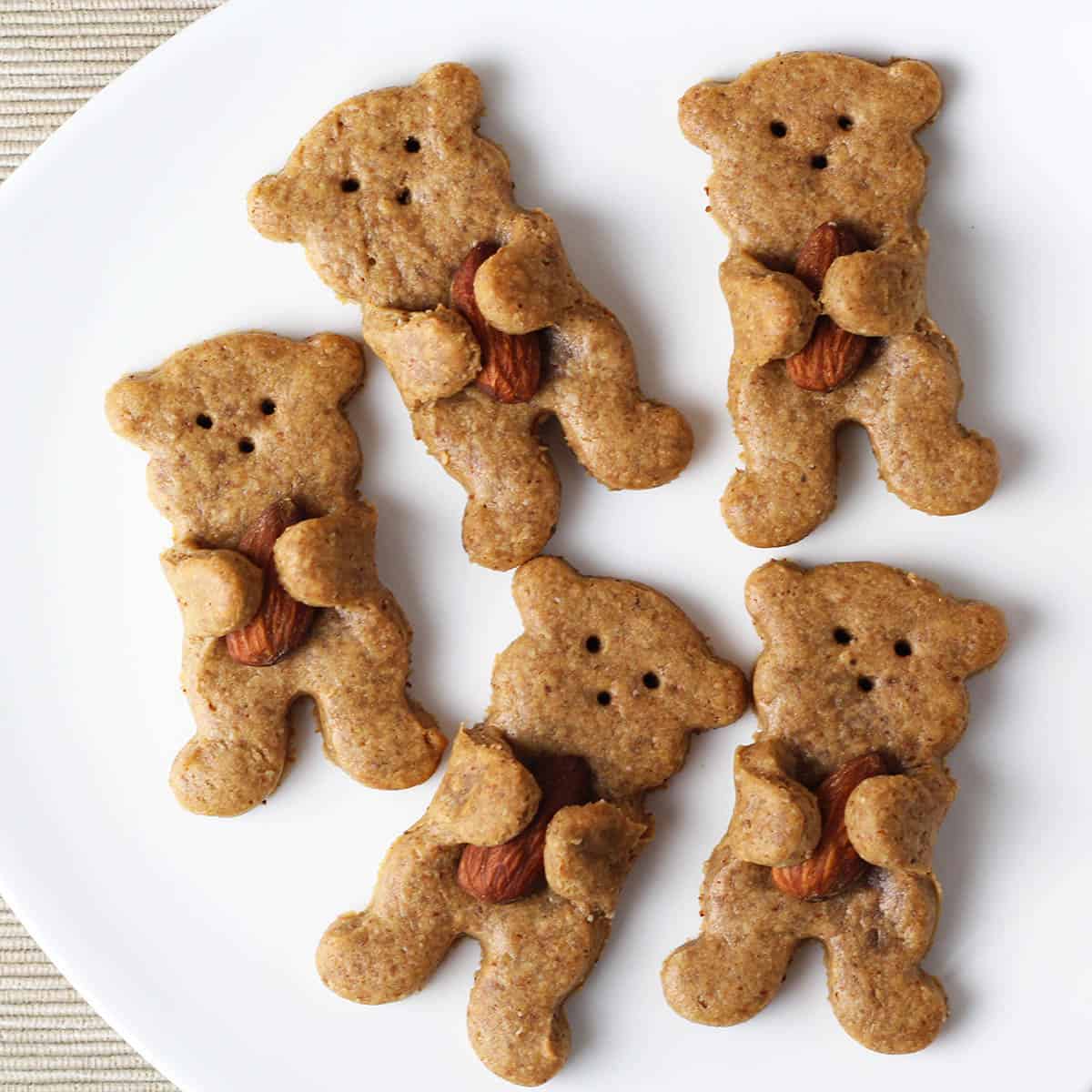 five small light brown bear shaped cookies that seem to be hugging almonds that are baked into them, on a white plate