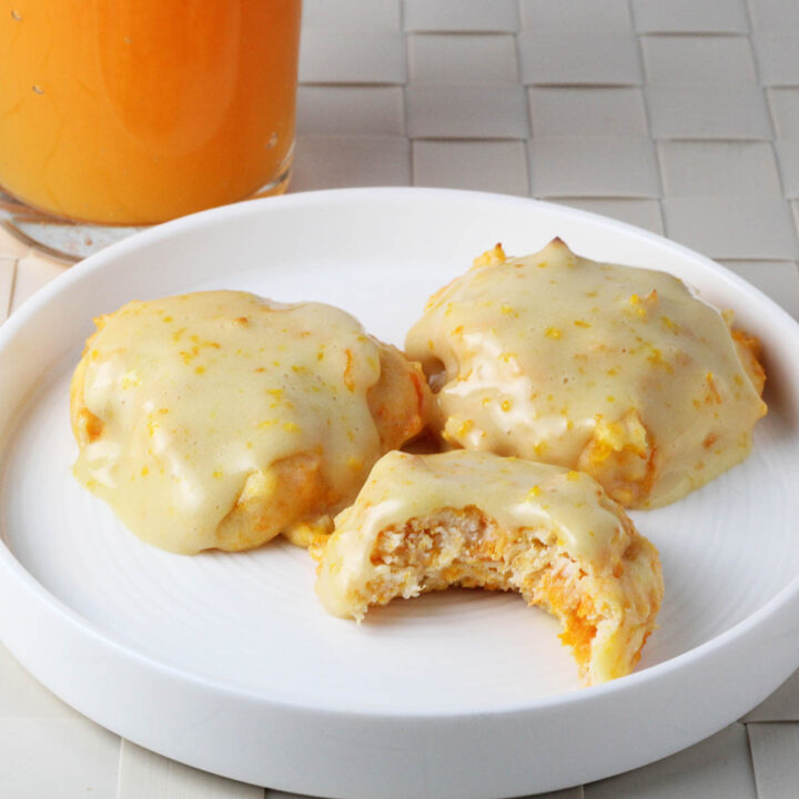 three orange iced carrot cookies on a white plate, one bitten, with a glass of orange carrot juice in the background