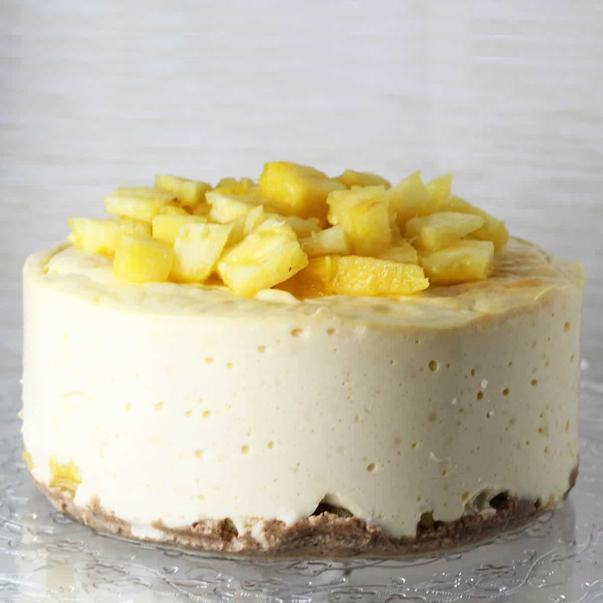 front view of a pineapple topped protein cheesecake on a flower etched glass cake plate with a light background