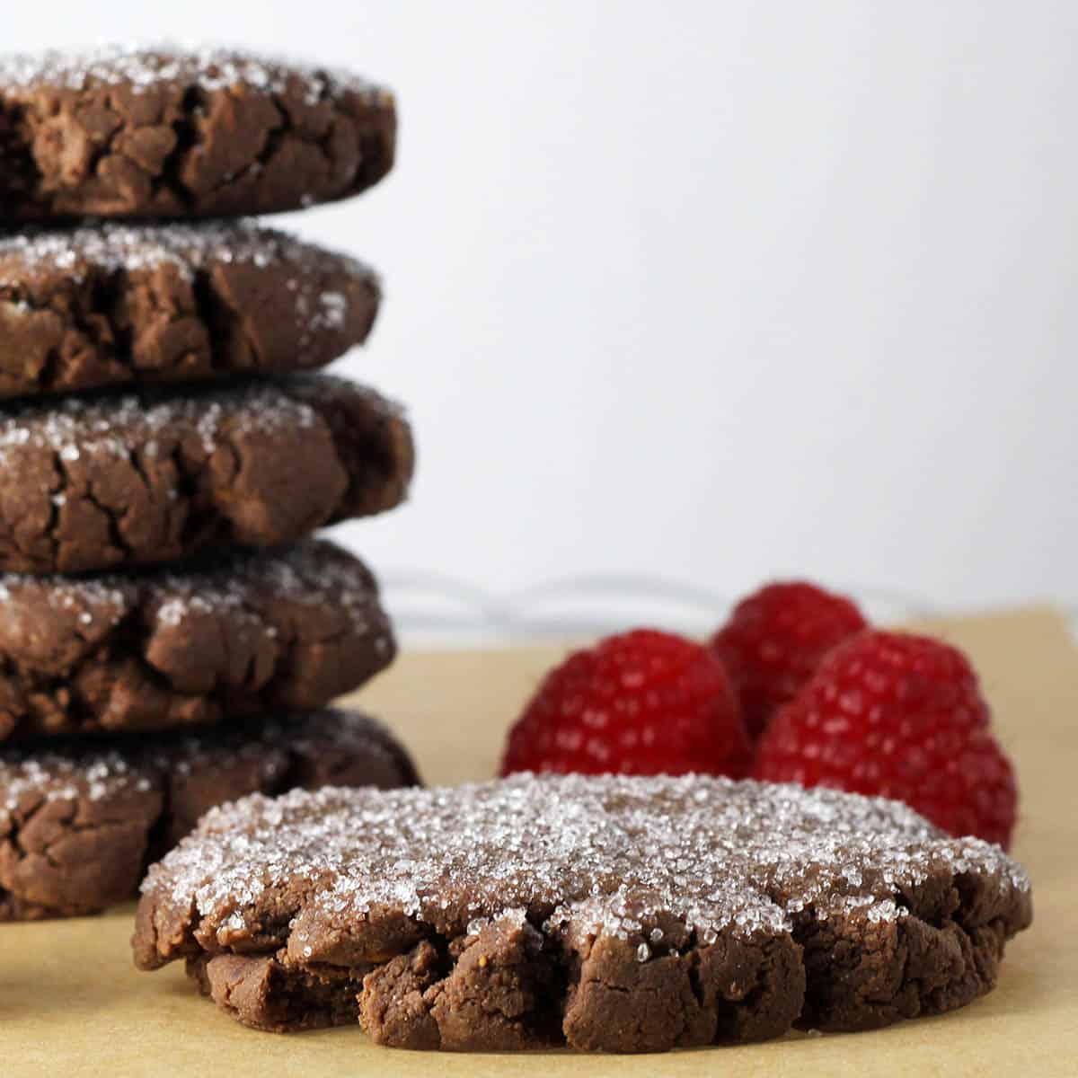 chocolate protein cookies topped with erythritol, five stacked in the back and one full cookie in the front with three fresh raspberries