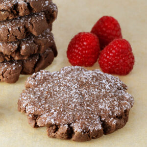 angled view of chocolate protein cookies topped with erythritol, five stacked in the back and one full cookie in the front with three fresh raspberries