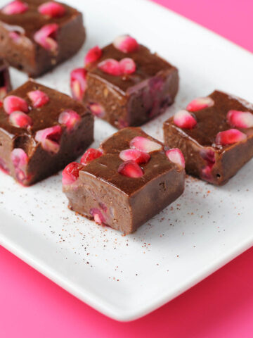 six pieces of chocolate protein fudge with pieces of pomegranate and a sprinkle of chili powder on a white plate on a hot pink background