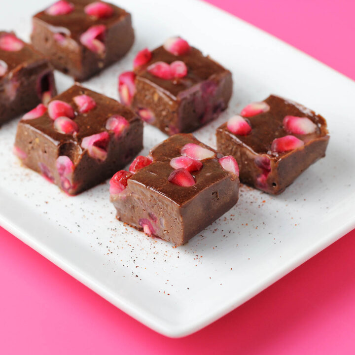 six pieces of chocolate protein fudge with pieces of pomegranate and a sprinkle of chili powder on a white plate on a hot pink background
